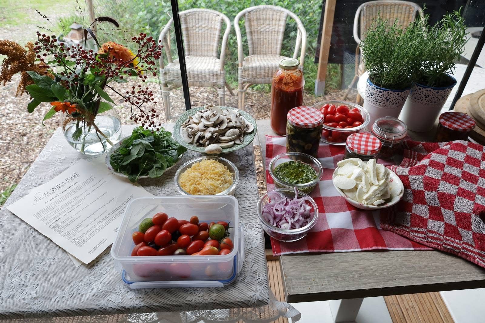 Tafel met schalen groenten, kruiden, tomaten en dipsauzen op een rood-wit geblokt kleed.