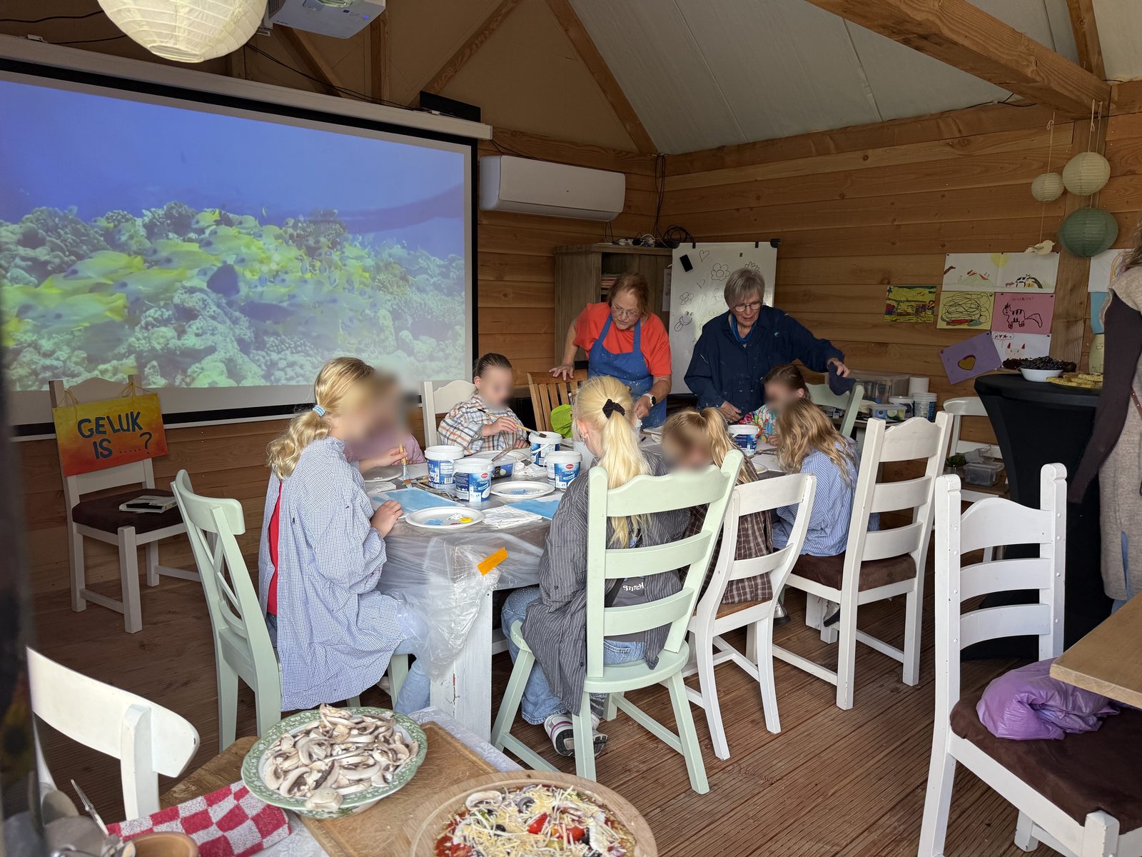 Binnenruimte met lange tafels waar kinderen en volwassenen schilderen; gezichten van kinderen zijn vervaagd.