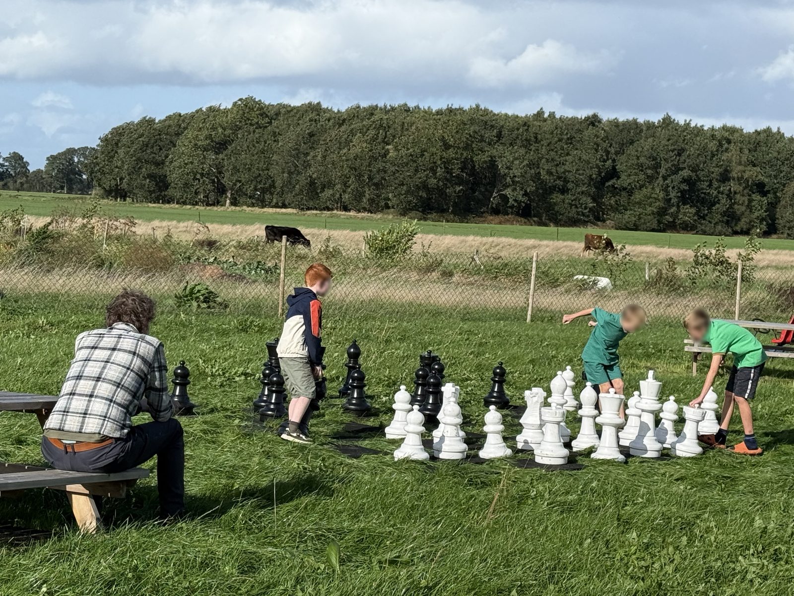 Groot zwart-wit schaakspel op het gras met spelende kinderen (onherkenbaar gemaakt) en een toekijkende volwassene.
