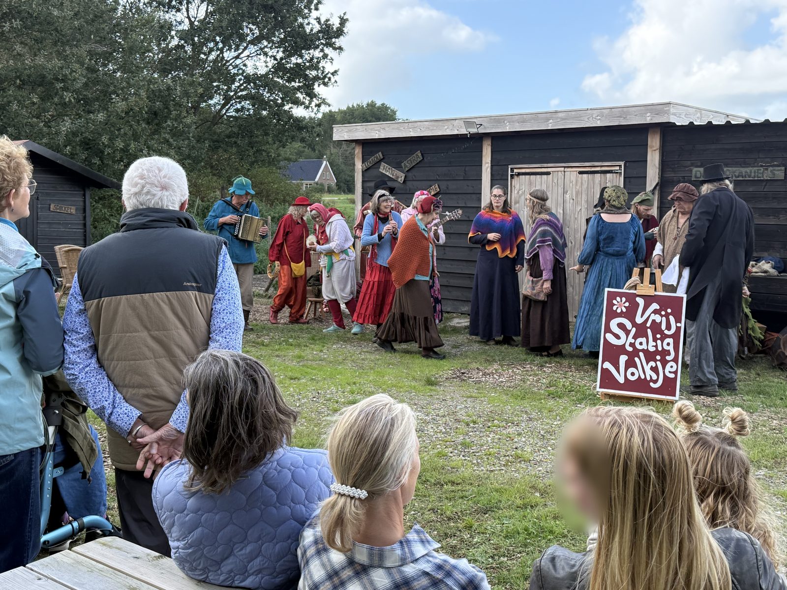 Spelers van het Vrij-Statig Volkje in kleurrijke kostuums treden op voor een publiek op het erf.