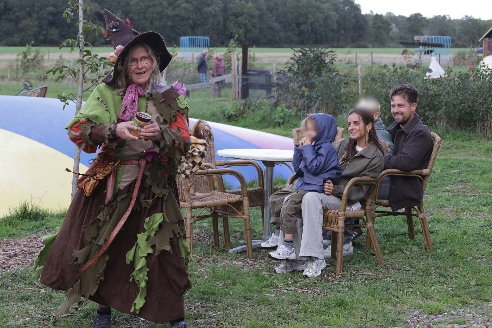 Verteller in een kostuum van bladeren staat op het gras; publiek zit op stoelen te luisteren.
