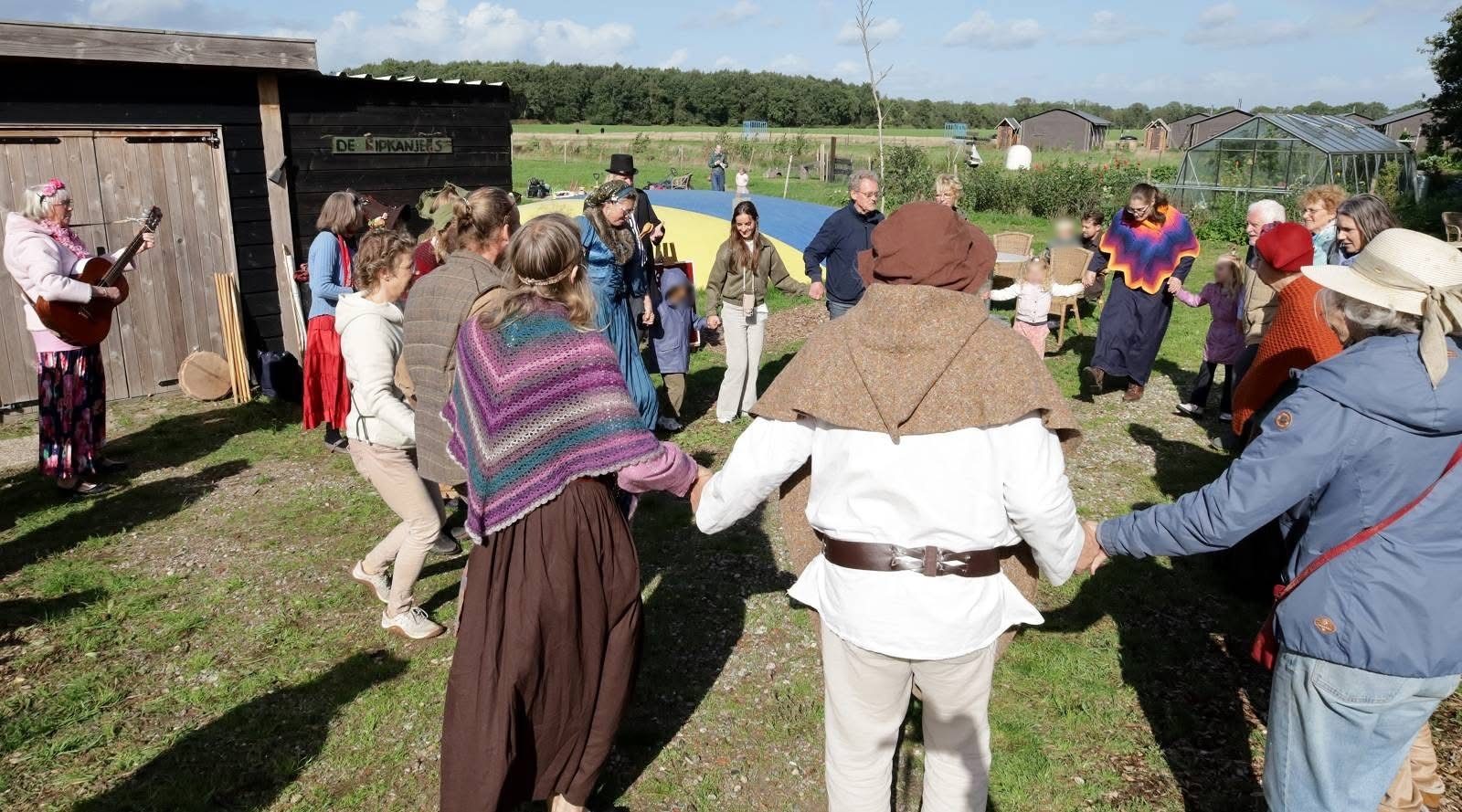Bezoekers en spelers dansen hand in hand in een kring op het gras bij de schuren.