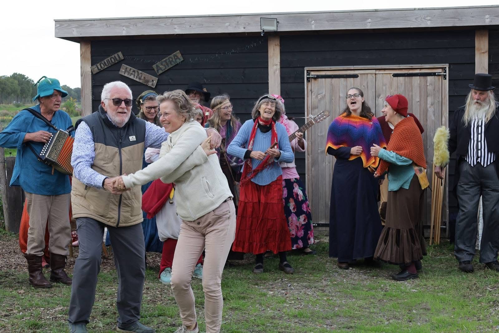 Een oudere man danst met een vrouw voor een rij spelers in kostuum bij de houten schuren.