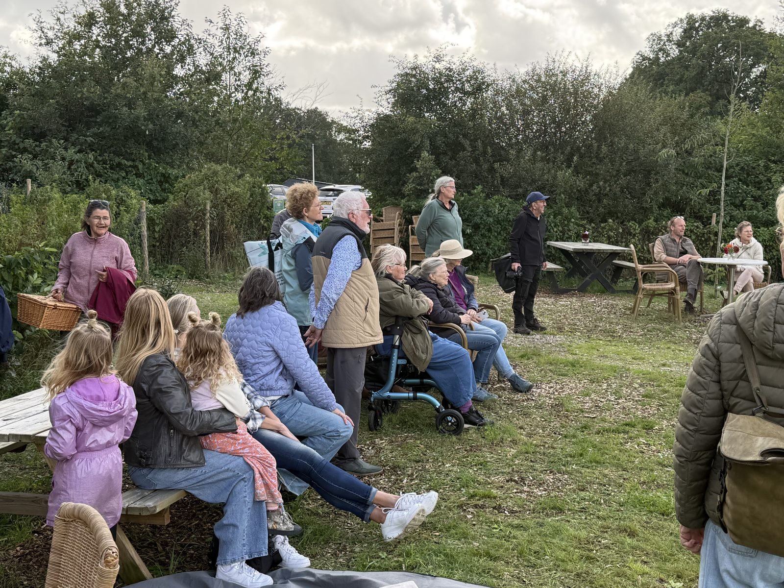 Publiek van alle leeftijden zit op stoelen en in het gras onder de bomen en kijkt naar de voorstelling; kinderen alleen van achteren te zien.