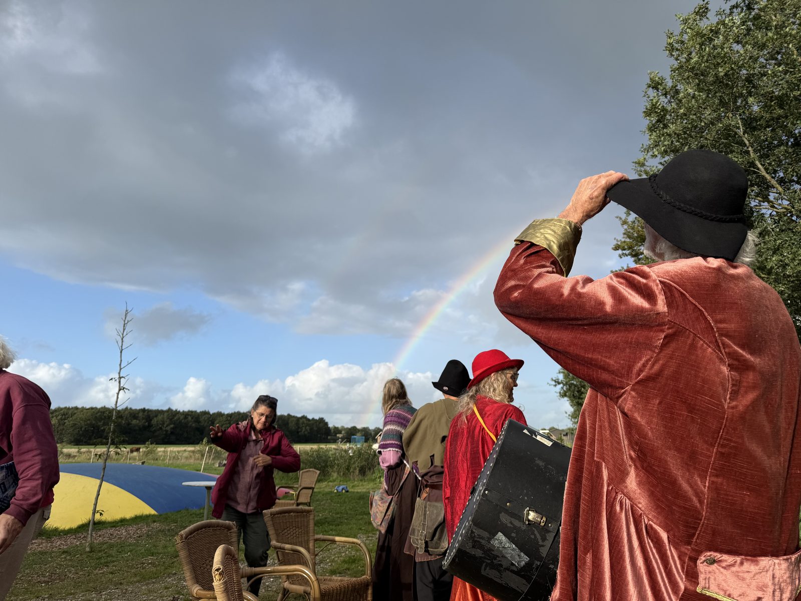 Regenboog boven het erf met op de voorgrond spelers in kostuum en een muzikant.